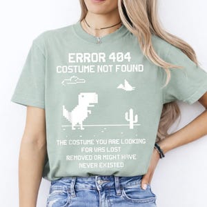 May include: A sage green short-sleeve t-shirt featuring a white pixelated graphic of a dinosaur, a cactus, and a cloud. The text reads: ERROR 404 COSTUME NOT FOUND. THE COSTUME YOU ARE LOOKING FOR WAS LOST REMOVED OR MIGHT HAVE NEVER EXISTED.