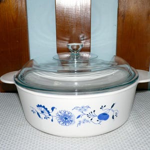 May include: A white round casserole dish with integrated side handles and a clear glass lid featuring a knob handle. The exterior of the dish is decorated with a blue floral pattern consisting of various stylized flowers, leaves, and vines.