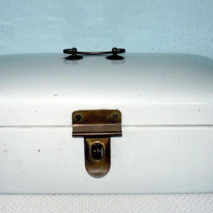 Peut inclure: Une boîte à pain rectangulaire blanche avec un dessus incurvé, un fermoir en laiton et une petite poignée. La boîte est en métal et a un aspect vintage. Conçue pour conserver le pain et les pâtisseries.
