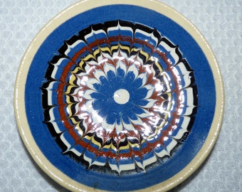 Könnte beinhalten: Ein runder Keramikteller mit einem blauen, weißen, schwarzen, gelben und braunen Blumenmuster. Der Teller hat eine blaue Mitte, einen weißen Rand und einen cremefarbenen Rand. Das Design ist symmetrisch und detailliert.