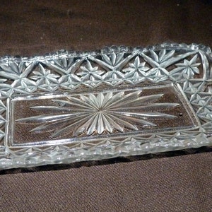 Peut inclure: Un plat rectangulaire en verre transparent avec un motif en forme d'éclat d'étoile au centre et des motifs en forme d'étoile sur les bords. Le plat a une bordure festonnée et est probablement utilisé pour servir ou exposer.