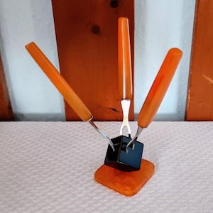 May include: A vintage fondue set with three orange-handled forks. The forks are set in a black square holder on an orange square base. The handles have a marbled appearance. The set is displayed on a white surface.