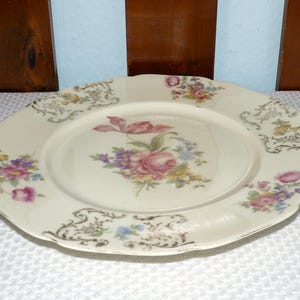 Könnte beinhalten: Cremefarbene Keramikplatte mit gewelltem Rand und Blumendesign. In der Mitte befindet sich eine rosa Rose und andere Blumen in Lila-, Gelb- und Blautönen. Dekorative goldfarbene Akzente umranden den Rand.