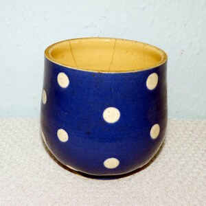 Könnte beinhalten: Eine blaue Keramiktasse mit weißen Punkten. Der Innenraum ist hellgelb. Die Tasse hat eine abgerundete Form und einen leicht unebenen Rand. Die Außenseite hat ein strukturiertes Aussehen.