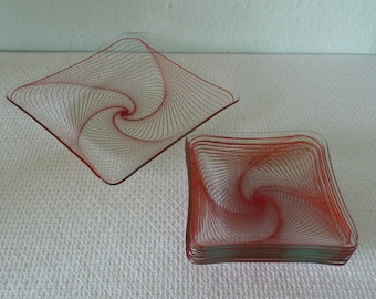 Könnte beinhalten: Ein Set aus quadratischen Glasplatten mit rotem Spiralmuster. Die Teller haben einen erhöhten Rand und ein einzigartiges Muster, das den Blick auf die Mitte lenkt. Die Teller sind gestapelt und auf einer weißen Oberfläche angeordnet.