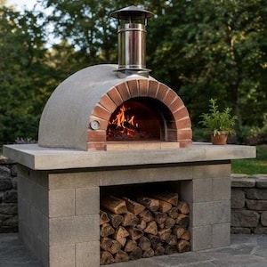 Può includere: Un forno per pizza da esterno a legna costruito in cemento grigio con un'apertura ad arco in mattoni rossi. Il forno è dotato di una canna fumaria in acciaio inossidabile, un termometro integrato e un vano inferiore con legna da ardere.
