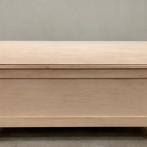 May include: A rectangular wooden storage chest featuring a smooth, light-colored natural wood finish. The chest has a prominent arched, dome-shaped lid and is supported by four carved, decorative feet at the base, showcasing a clean, unfinished look.