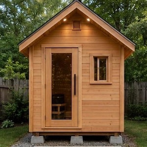 May include: A small, detached wooden sauna cabin with horizontal cedar siding, a gabled roof, and a single glass-panelled door with a long black handle. The structure features a small square window, two recessed ceiling lights, and a vent.