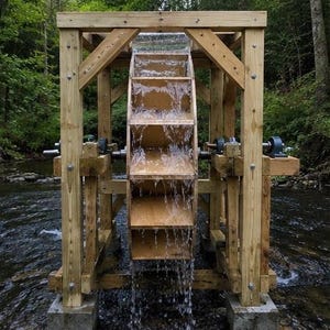 May include: A wooden water wheel mounted within a sturdy timber frame structure, positioned over a flowing stream. The wheel features multiple rectangular paddles with water cascading over them, supported by metal bearings on a horizontal axle.