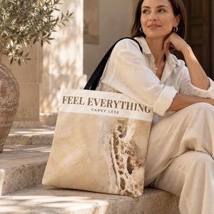 May include: A white tote bag with black shoulder straps featuring the text FEEL EVERYTHING CARRY LESS in black lettering. The lower portion of the bag displays a detailed, textured beige and brown rock formation graphic print.