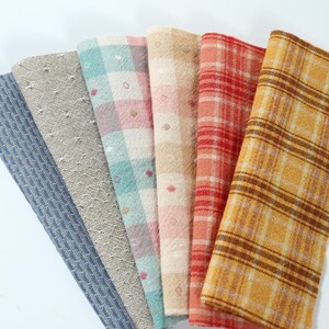 May include: A collection of six folded fabric swatches arranged in a fan shape. The patterns include a blue herringbone, a beige dotted texture, a pastel plaid, a beige grid, a red plaid, and a yellow plaid with brown stripes.