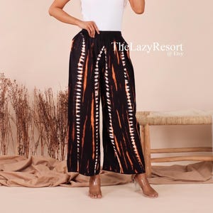 May include: A white short-sleeved t-shirt paired with black wide-leg trousers featuring a vertical orange and white tie-dye pattern. The outfit includes a wide-brimmed beige sun hat and clear heeled sandals. The text TheLazyResort @ Etsy is visible.