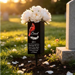 Pode incluir: Uma estaca memorial retangular preta com um pássaro cardeal vermelho, uma pena branca e o texto "Dad" seguido de "Gone Yet Not Forgotten, Although We Are Apart, Your Spirit Lives Within Me, Forever In My Heart." Peônias brancas preenchem o topo.