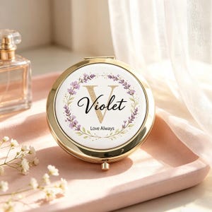 Może przedstawiać: Okrągłe, złote lusterko kieszonkowe z białym wieczkiem ozdobionym złotą, brokatową literą V, imieniem Violet napisanym czarną kursywą oraz wieńcem z kwiatów lawendy. Poniżej imienia znajduje się czarny napis Love Always.
