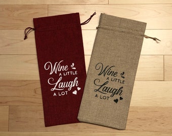 May include: Two wine bottle gift bags made of burlap fabric. One bag is red and the other is beige. Both bags have the text "Wine a little laugh a lot" printed on them in white and black.