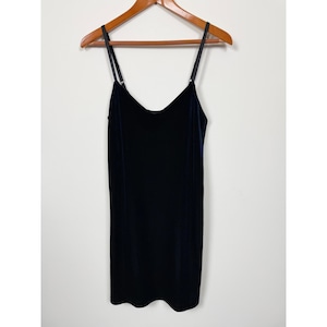 May include: A dark navy blue velvet slip dress featuring a V-neckline and thin, adjustable black spaghetti straps. The garment has a straight, simple silhouette and is displayed on a wooden hanger against a plain, light-colored background.