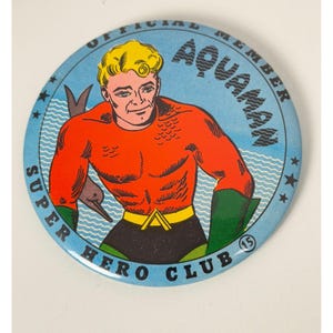 Puede incluir: Una chapa redonda con una ilustración de cómic de Aquaman con pelo rubio, camisa naranja y guantes verdes. El fondo azul incluye líneas onduladas y el texto OFFICIAL MEMBER AQUAMAN SUPER HERO CLUB 15.