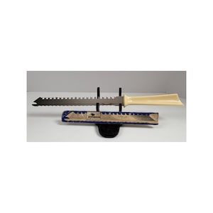 May include: A serrated metal knife with a cream-colored handle, positioned horizontally on a black stand. Below the blade rests a rectangular cardboard sleeve featuring blue edges and a central label with the text "EKCO FLINT" and "USA".