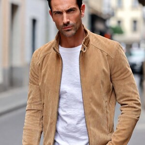 Könnte beinhalten: Eine hellbraune Wildlederjacke mit Stehkragen, durchgehendem Frontreißverschluss und langen Ärmeln, getragen über einem schlichten weißen T-Shirt und einer dunklen Hose. Die Jacke zeichnet sich durch eine glatte Textur und strukturierte Schultern aus.