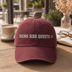 Könnte beinhalten: Eine kastanienbraune Baseballkappe mit dem gestickten weißen Text DOING SIDE QUESTS, gefolgt von einem kleinen Symbol eines zwanzigseitigen Würfels. Die Kappe hat eine strukturierte Krone, einen gebogenen Schirm und sichtbare Nähte.
