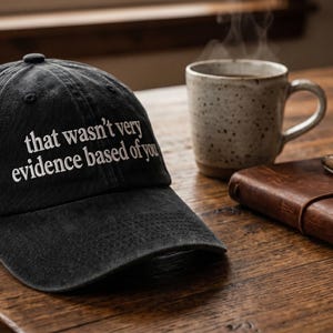 Könnte beinhalten: Eine anthrazitfarbene Baseballkappe mit dem weiß gestickten Text "that wasn't very evidence based of you". Die Kappe hat eine strukturierte Krone, einen gebogenen Schirm und einen Knopf an der Oberseite im klassischen Sechs-Panel-Design.
