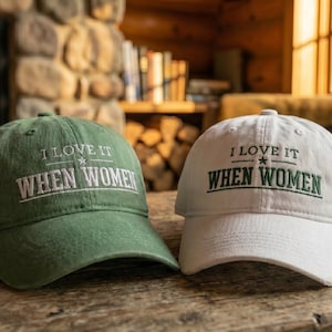 Könnte beinhalten: Zwei Baseballkappen, eine olivgrün und eine weiß, jeweils mit gesticktem Text I LOVE IT WHEN WOMEN in einer Serifenschrift und einem kleinen Stern, der zwischen den beiden Textzeilen auf der Vorderseite der Kappen zentriert ist.