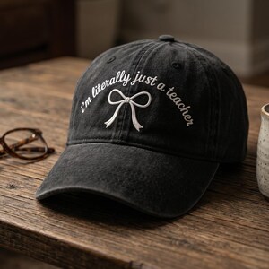 May include: A black baseball cap featuring white embroidered text that reads I'm literally just a teacher with a white bow graphic centred below the text. The cap has a curved brim and a structured crown with a button on the top centre.