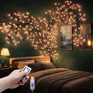 Op de afbeelding: Een aan de muur gemonteerde LED-kersenbloesemboomlamp met donkere takken en gloeiende roze bloemvormige lampjes. Het licht wordt bediend met een afstandsbediening en een USB-aansluiting. De decoratie is geplaatst boven een bed in een slaapkamer.