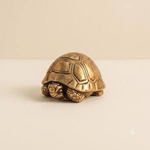 Peut inclure: Une petite figurine de tortue en laiton massif avec une finition dorée métallique polie. La sculpture présente un motif de carapace segmenté détaillé et une peau texturée sur la tête et les pattes, la tortue étant en position rétractée.