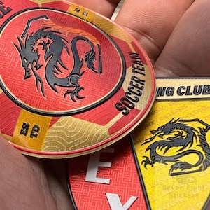 May include: Two circular patches featuring black dragon silhouettes. The top patch is red and gold with the text SOCCER TEAM and ESTD. The bottom patch is yellow and black with the text ING CLUB and Seven Eight Stickers visible in the background.