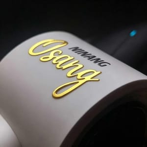 May include: A white cylindrical object featuring the word Osang in a raised, metallic gold script font, positioned above the word NINANG printed in a smaller, black, sans-serif typeface. The surface is smooth and curved with a matte white finish.