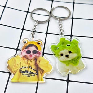 Puede incluir: Dos llaveros de acrílico transparente con anillas y cadenas de metal. Uno muestra a una persona con sudadera amarilla con capucha y el texto "adore Farense badblaod INSPIRTION FONT". El otro muestra un perro blanco con capucha verde.