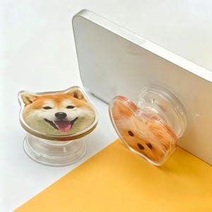 May include: Two circular phone grips with clear plastic bases. One grip features a printed image of a Shiba Inu dog with a smiling expression, while the other grip is attached to a white smartphone and displays a close-up image of a small dog face.