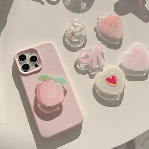 May include: A light pink smartphone case with a peach-shaped phone grip attached. Five additional phone grips are arranged nearby, featuring designs of a white flower, a pink strawberry, a pink ribbon, a heart with the word Love, and a heart.