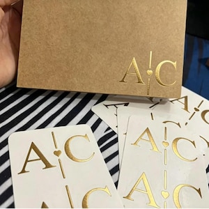 May include: A rectangular tan cardboard box and several white rectangular stickers, each featuring the gold foil-stamped letters A and C separated by a vertical line with a small heart icon in the center, creating a matching monogram design.