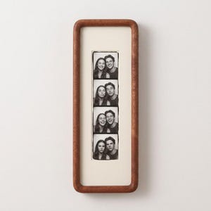 May include: A rectangular wooden photo frame with a cream mount. The frame holds four black and white photo booth strips, each featuring a couple. The frame has rounded edges and a warm brown finish, ideal for displaying memories.