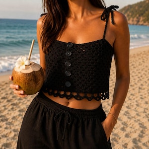 May include: A black crochet crop top featuring thin shoulder straps, a square neckline, and a vertical row of five black buttons down the center. The hem is finished with a scalloped crochet pattern, and the garment is paired with black pants.