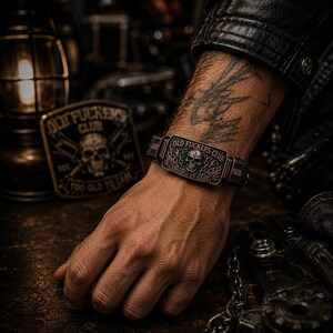 May include: A dark metal bracelet featuring a rectangular plate with a central skull graphic, flame details, and the text "OLD FUCKERS CLUB" and "TOO OLD TO CARE" engraved on the surface. The bracelet has a textured band and a weathered finish.