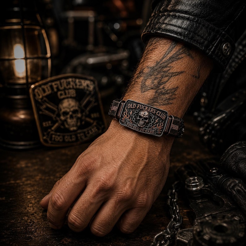 May include: A dark metal bracelet featuring a rectangular plate with a central skull graphic, flame details, and the text "OLD FUCKERS CLUB" and "TOO OLD TO CARE" engraved on the surface. The bracelet has a textured band and a weathered finish.