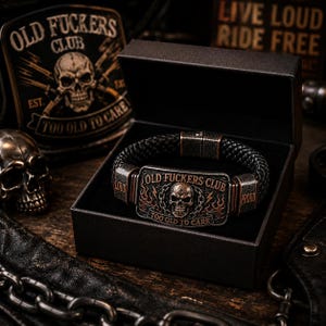May include: A black braided leather bracelet featuring a rectangular metal plate with a skull graphic, flames, and the text OLD FUCKERS CLUB and TOO OLD TO CARE. The bracelet has metal side accents labeled LOUD and PROUD, presented in a black box.