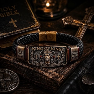 May include: A black braided leather bracelet featuring a rectangular antique brass-toned metal plate. The plate displays a relief of a crowned figure, the text KING OF KINGS, CHRIST IS MY STRENGTH, JESUS SAVES, and GOD IS WITH ME.