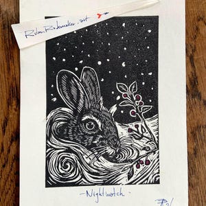 Op de afbeelding: Een zwart-witte linosnede op papier met een konijn omringd door wervelende patronen en een takje met rode bessen tegen een sterrenhemel. De prent is getiteld Nightwatch en gesigneerd met R26, inclusief een papieren label met Rulen Rodemaker.art.