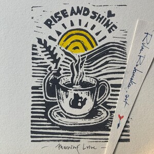 Op de afbeelding: Een zwart-gele linosnede met een dampende kop koffie op een schoteltje, een klein vaasje met een varen en een zon. De tekst luidt RISE AND SHINE bovenaan en Morning Love onderaan. Een strook papier met de tekst Rulan Rosenker.art is zichtbaar.