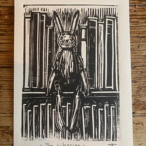 Op de afbeelding: Zwart-wit linoleumsnede van een konijnfiguur voor een boekenplank. Het konijn heeft lange oren en draagt een jurk. De tekst "- The Librarian -" staat onder de afbeelding.