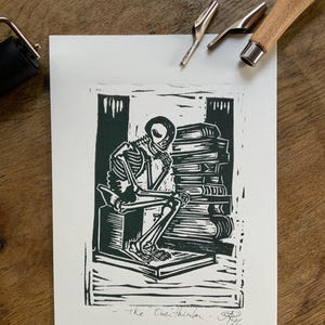 Op de afbeelding: Een zwart-witte linosnede op wit papier met een skelet dat op een blok zit, de kin op een hand rustend terwijl het tegen een hoge stapel boeken leunt. De tekst "The Overthinker" is onderaan in het midden met potlood handgeschreven.