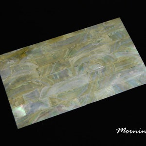 May include: A close-up of a decorative tile made of iridescent mother-of-pearl shell. The tile is rectangular and is made up of many small, irregular pieces of shell arranged in a pattern. The shell has a light green and white colour with a subtle shimmer.