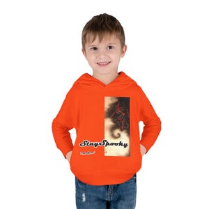 Puede incluir: Una sudadera con capucha de color naranja brillante que presenta un estampado gráfico rectangular en la parte delantera. El gráfico muestra un diseño artístico oscuro y arremolinado con el texto "Stay Spooky" en negrita y "PeaceSnow" debajo.