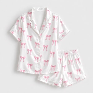 Könnte beinhalten: Ein weißes Pyjama-Set bestehend aus einem kurzärmeligen Hemd mit Knopfleiste und Reverskragen sowie passenden Shorts. Der Stoff ist mit einem durchgehenden Muster aus kleinen, hellrosa Schleifen bedruckt. Das Hemd hat vier weiße Knöpfe.