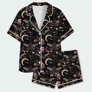May include: A black short-sleeve pajama set with white piping, featuring a repeating pattern of moths, crescent moons, and pink flowers. The button-down shirt has a notched collar, and the matching shorts have an elasticized waistband.