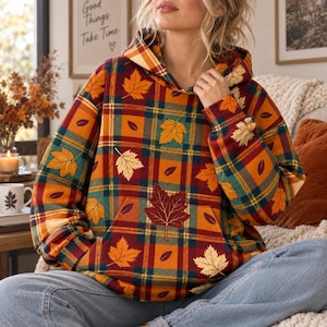 May include: A long-sleeve hooded sweatshirt featuring a plaid pattern in shades of red, orange, yellow, and teal. The garment is decorated with a repeating print of various maple leaves in gold, orange, and dark red colors, and includes a front pocket.